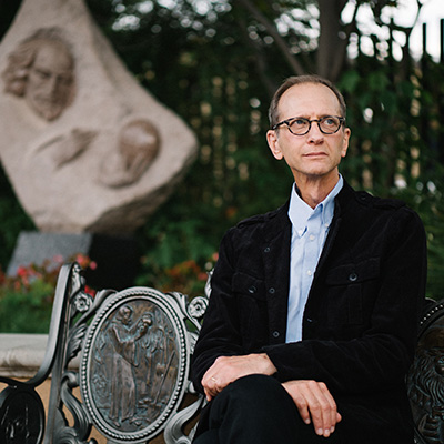 Playwright Will Cooper in black jacket, blue shirt, and glasses sitting on a bench 